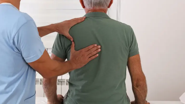 A clinician placing both hands on an older man upper back during a seated examination