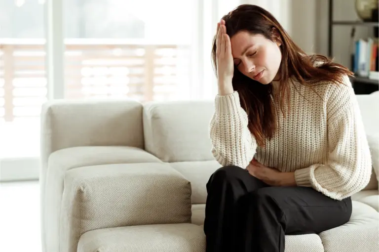 Woman sitting on a sofa with one hand to her head, worn down by ongoing pain