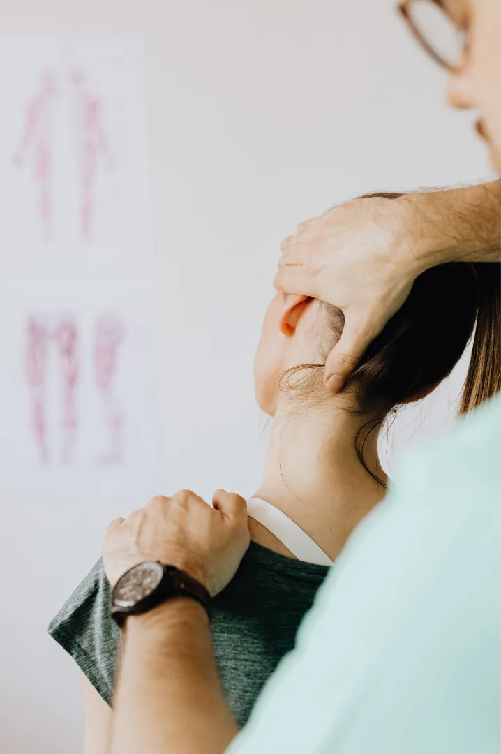 A clinician supporting a patient head and shoulder during a neck assessment, with anatomy charts on the wall