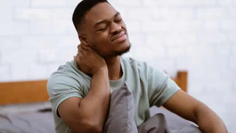 A man sitting on a bed holding the side of his neck