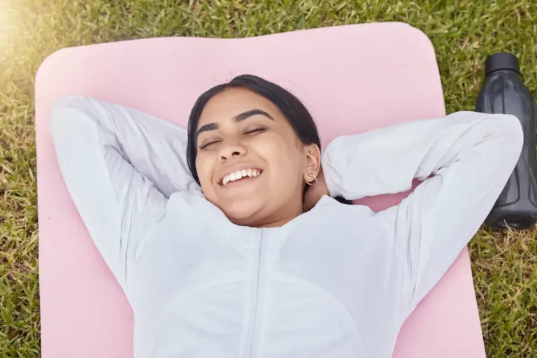 A woman lying on an exercise mat on grass with her hands behind her head, resting after a workout